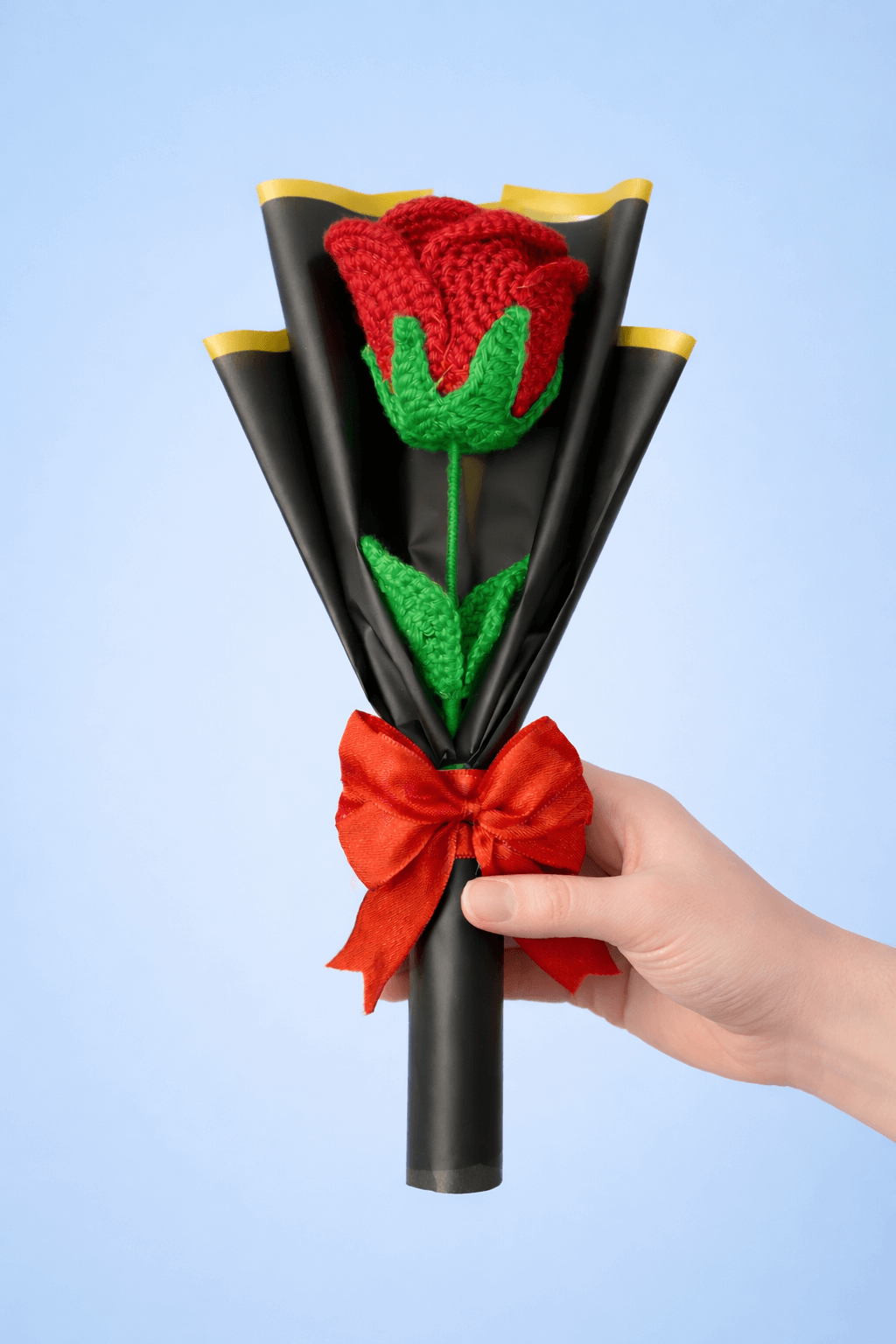 Hand holding a bouquet made of black paper, red ribbon, and green leaves against a light blue background