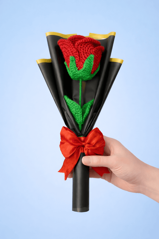 Hand holding a bouquet made of black paper, red ribbon, and green leaves against a light blue background