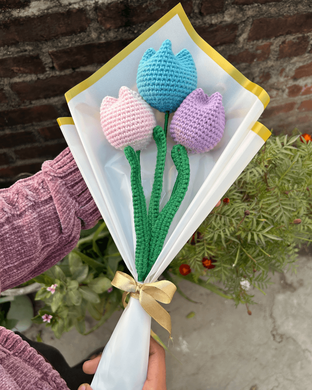 Crocheted tulips in blue, pink, and purple held against a brick wall.